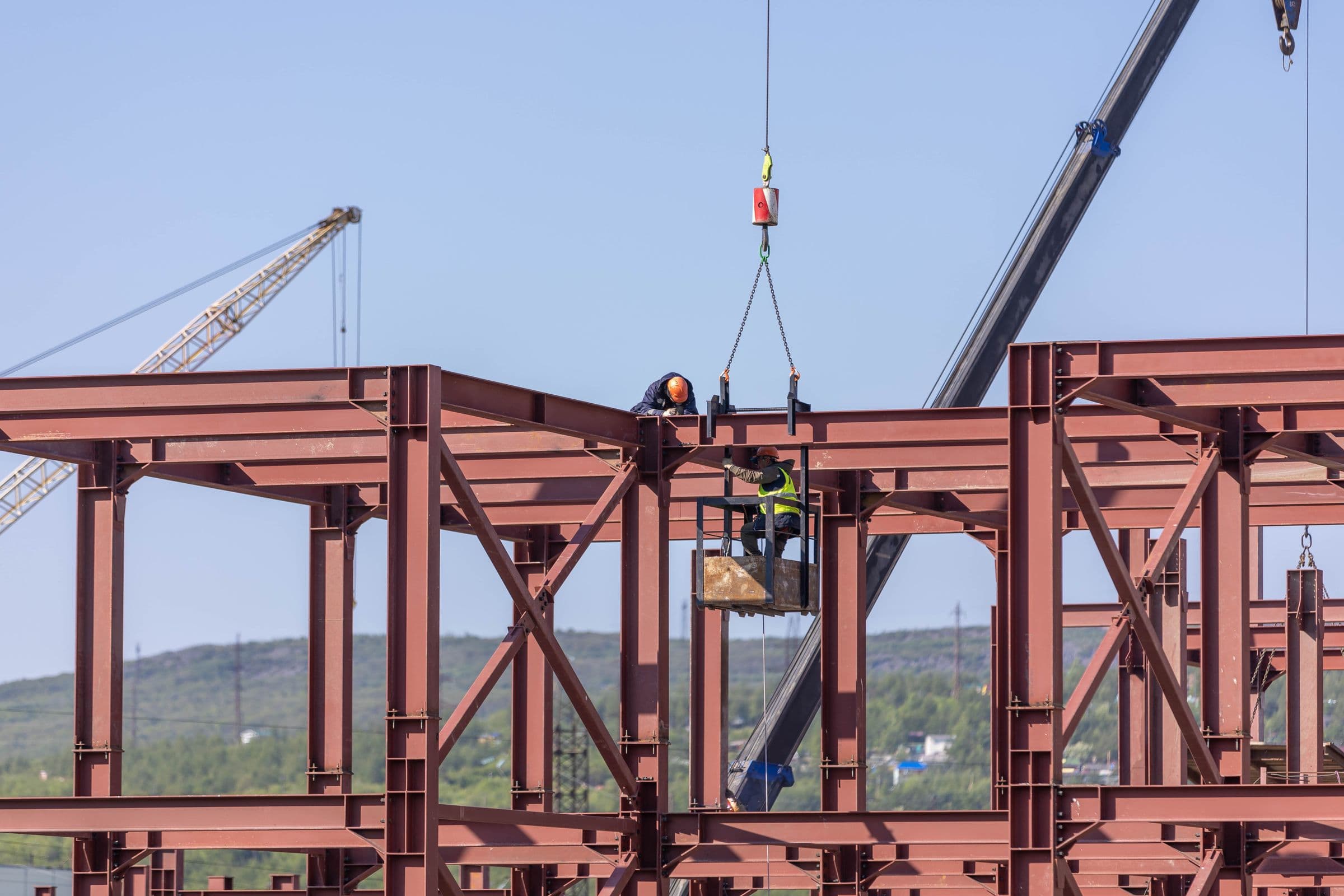 Two construction workers guide a steel member into an active structural frame